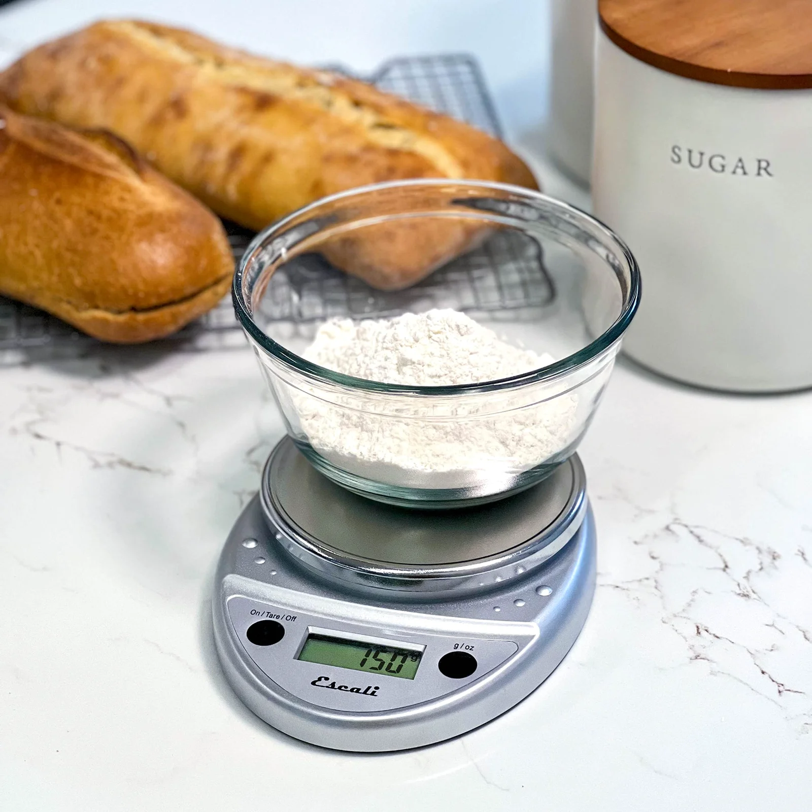 A loaf and a bowl of flour being weighed on a chrome Escali Primo scale on a marble counter